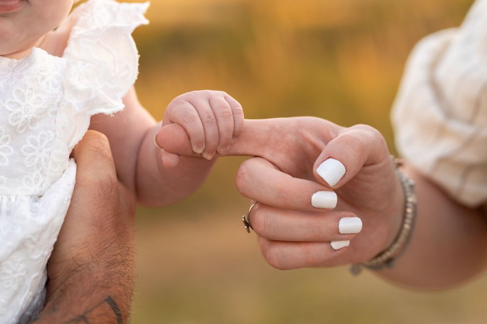 A baby grasping an adult's finger with white manicured nails and bracelets, held by another adult outdoors.