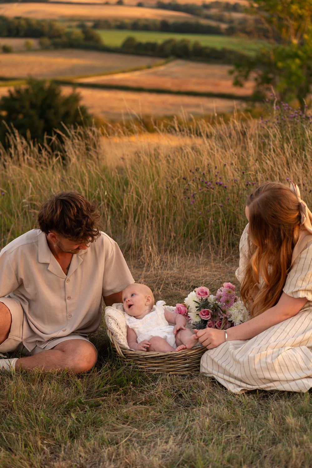 A couple sits in a golden field at sunset, lovingly gazing at their newborn baby in a wicker basket with flowers.