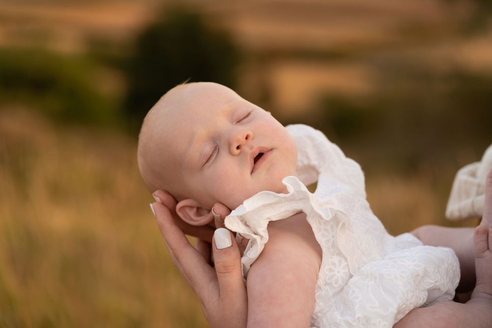 Peaceful sleeping newborn baby in white lace outfit cradled in gentle hands outdoors in golden field.