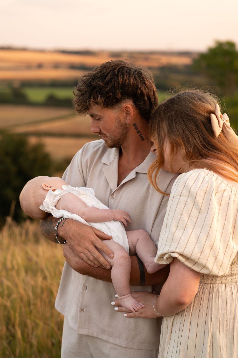 Young parents holding their newborn baby in a golden field at sunset, wearing neutral linen outfits.