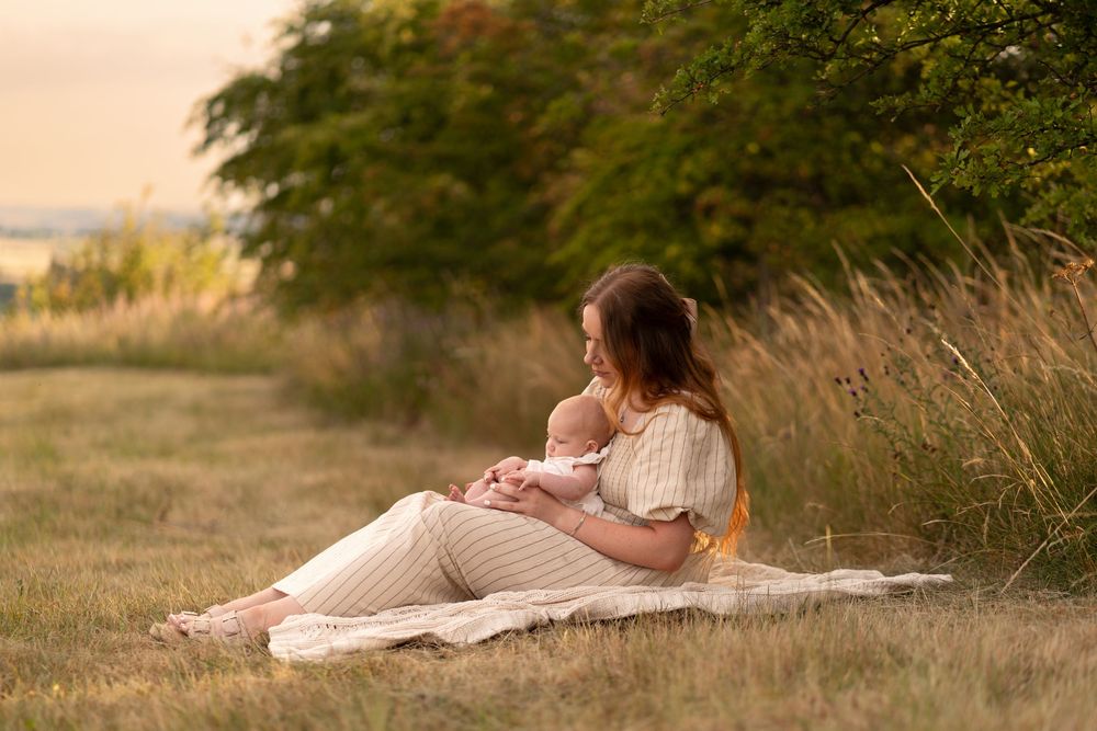 Mother breastfeeding newborn baby outdoors on blanket in golden field at sunset.
