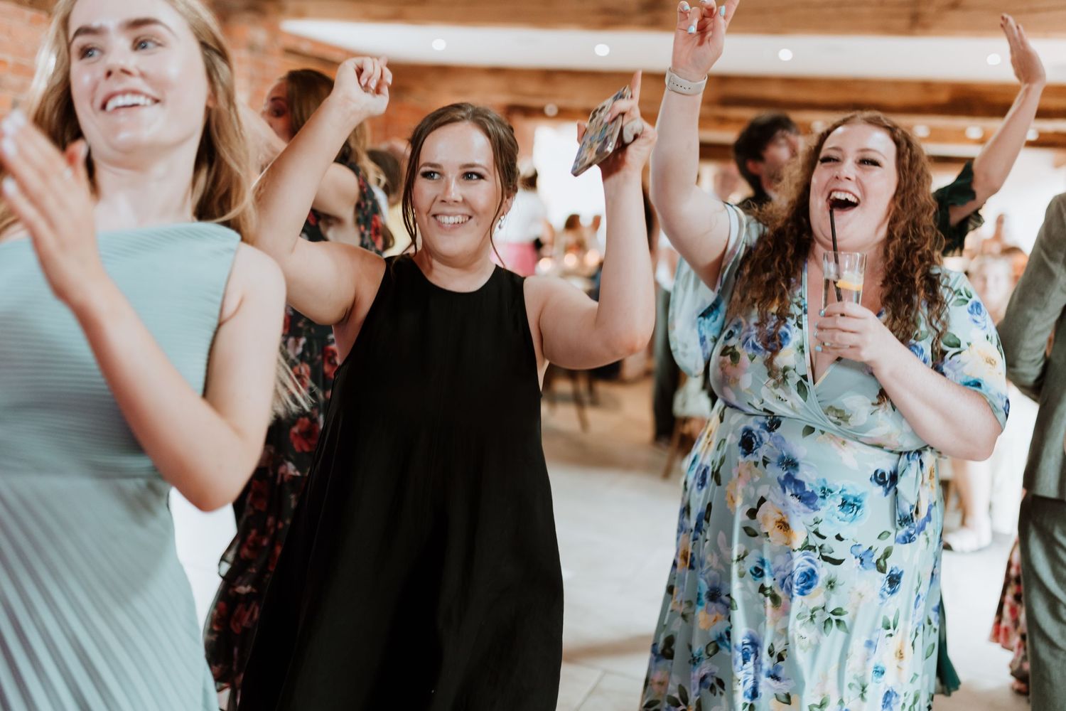 Three joyful women dancing and celebrating at a wedding reception in a rustic venue.