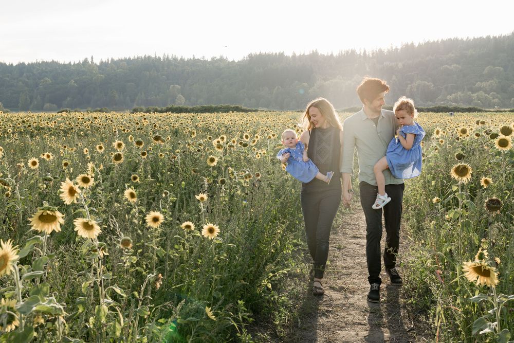 Family walking through sunflower field at sunset, parents carrying two young children in blue outfits.