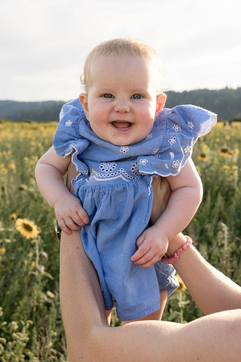 Smiling baby in blue dress held up by adult hands in a sunny sunflower field.