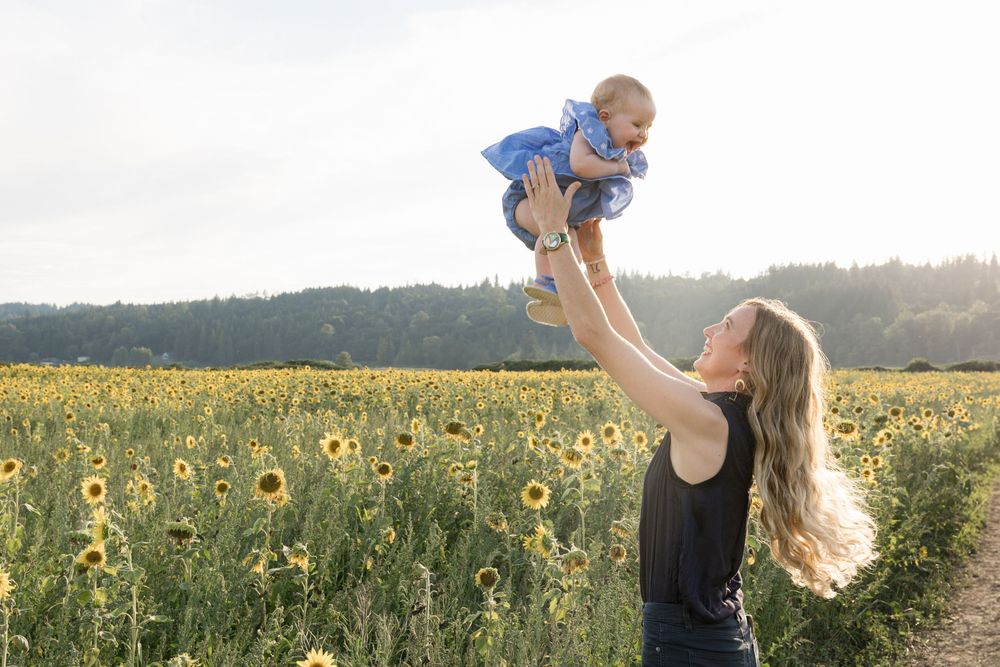 A mother joyfully lifts her baby in the air in a vibrant sunflower field on a sunny day.