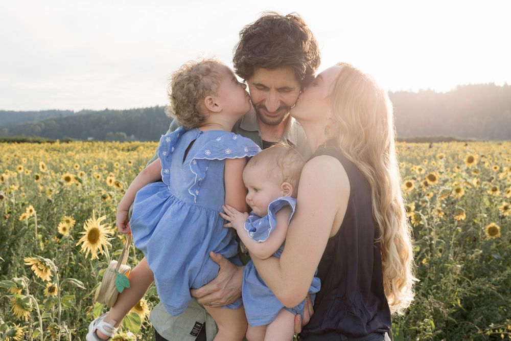 Family of four posing in a sunflower field at golden hour, parents kissing children in matching blue outfits.