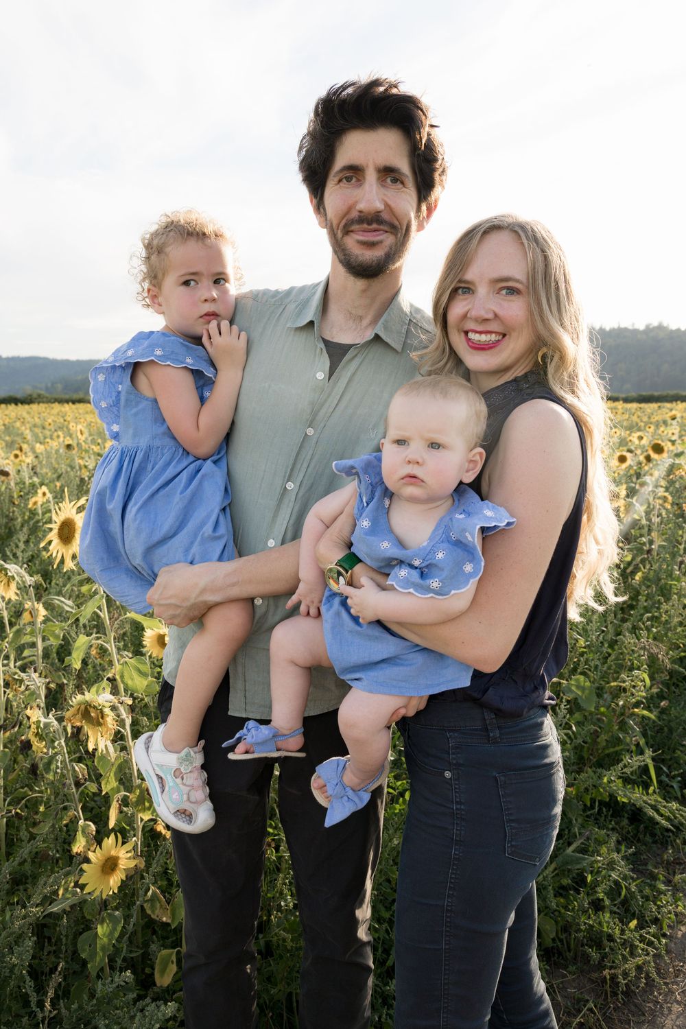 A happy family of four poses in a sunflower field, parents holding two young girls in matching blue dresses.