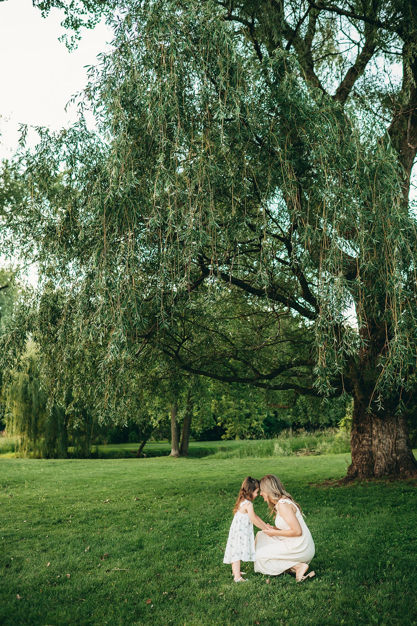 Mother and young daughter in white dresses share a tender moment under a weeping willow tree on lush green grass.