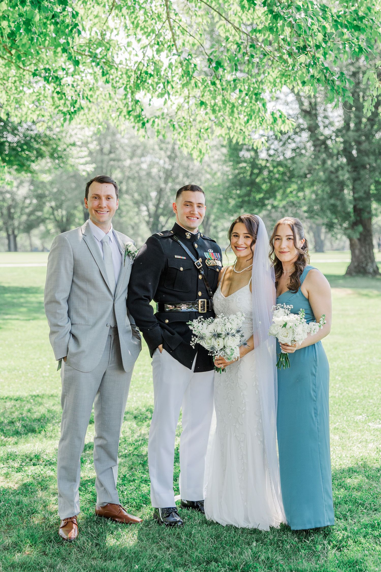 Wedding party poses outdoors, featuring a groom in military dress uniform, bride in white gown, groomsman, and bridesmaid in blue.