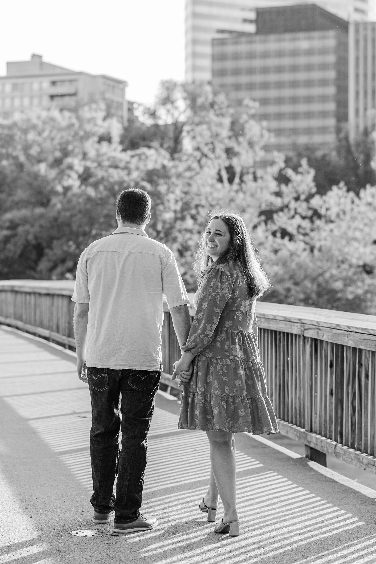 Black and white photo of a couple holding hands on a bridge, woman smiling back at camera with city skyline behind.