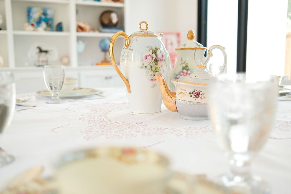 Elegant tea party table setting with white and gold teapots, fine china plates, and crystal glasses on a white tablecloth.