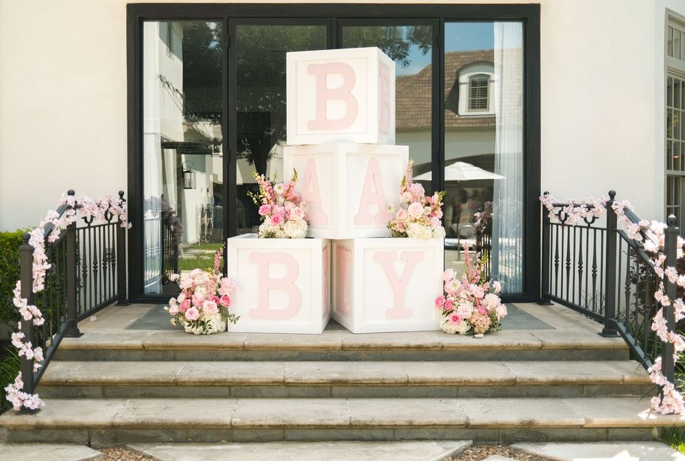 Pink baby shower decor with large letter blocks spelling BABY, floral arrangements, on decorated steps.