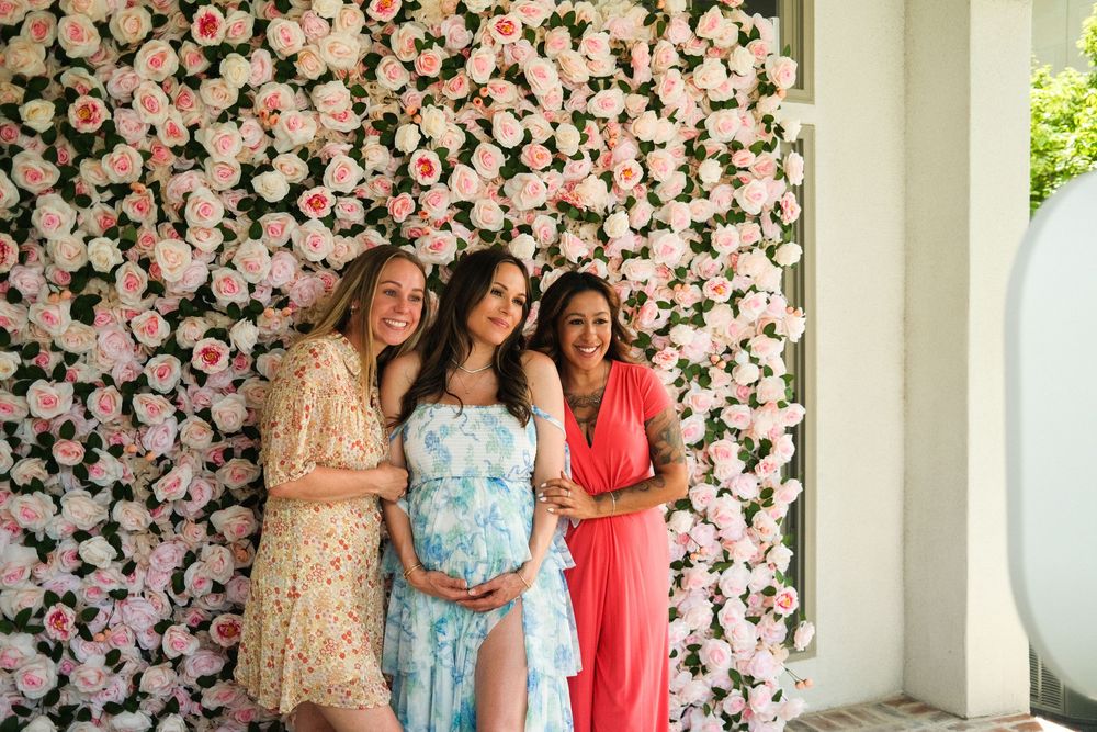 Three women pose smiling in front of a pink floral wall, with the middle woman cradling a baby bump.