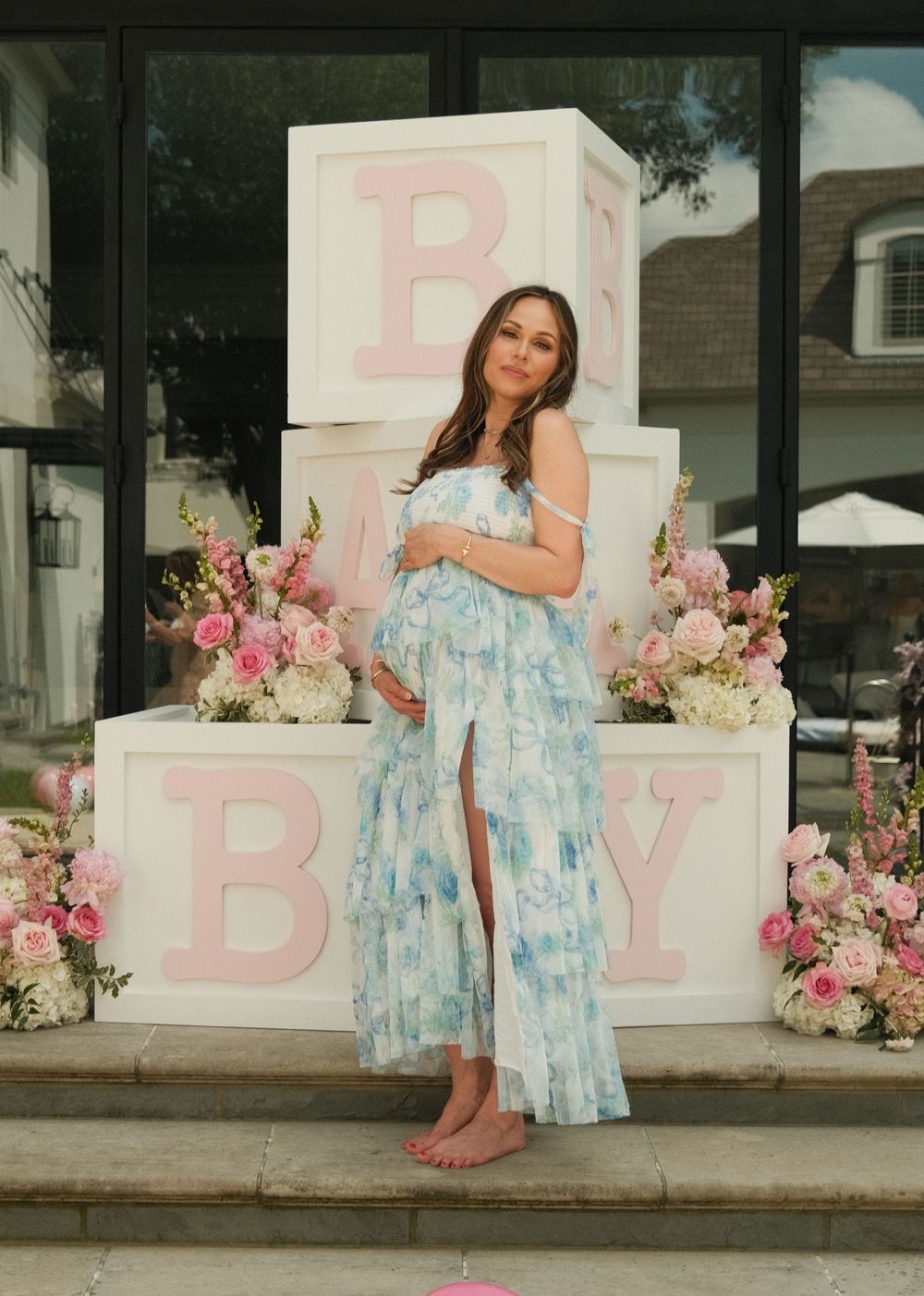 Pregnant woman in floral blue maxi dress poses at baby shower with large white BABY blocks and pink flowers.