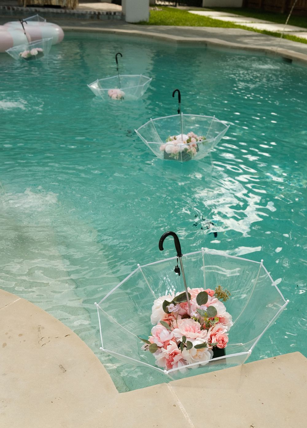 Clear umbrellas with pink flowers floating in a turquoise swimming pool, creating an elegant decorative display.