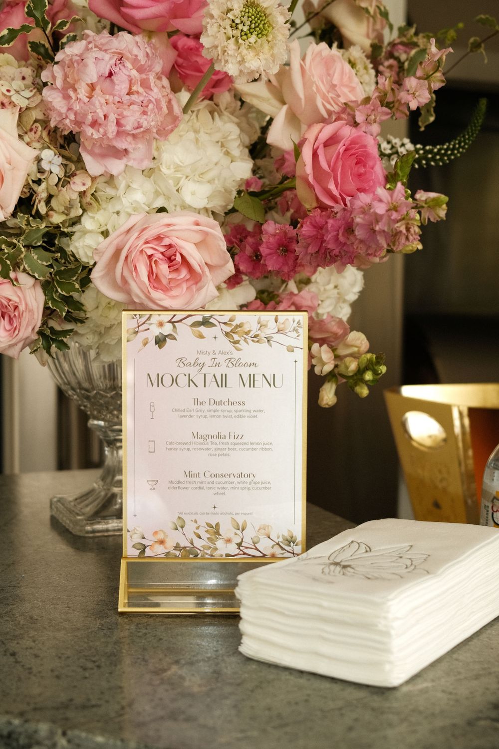 Elegant mocktail menu display with gold frame beside lush pink floral arrangement at a baby shower event.