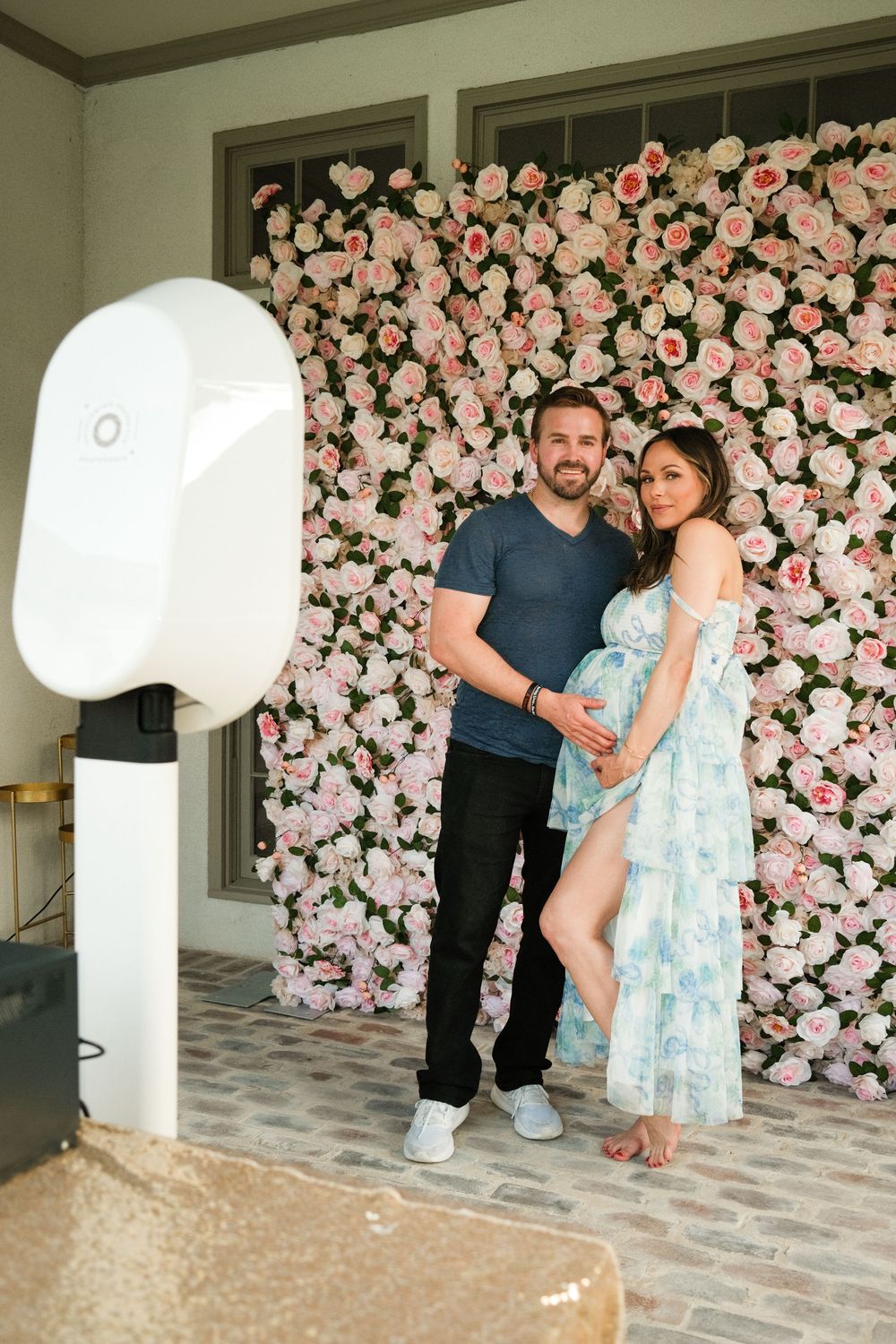 Pregnant couple posing in front of a pink floral wall backdrop at a baby shower event.