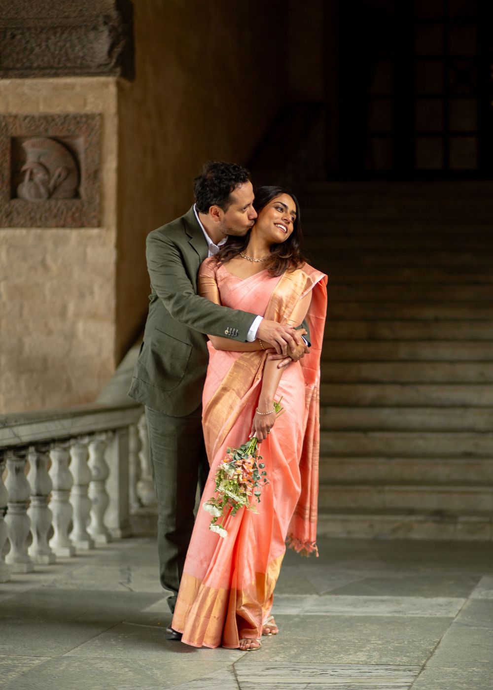 Couple in formal attire, woman in pink saree holding bouquet, man in suit embracing her on ornate staircase.