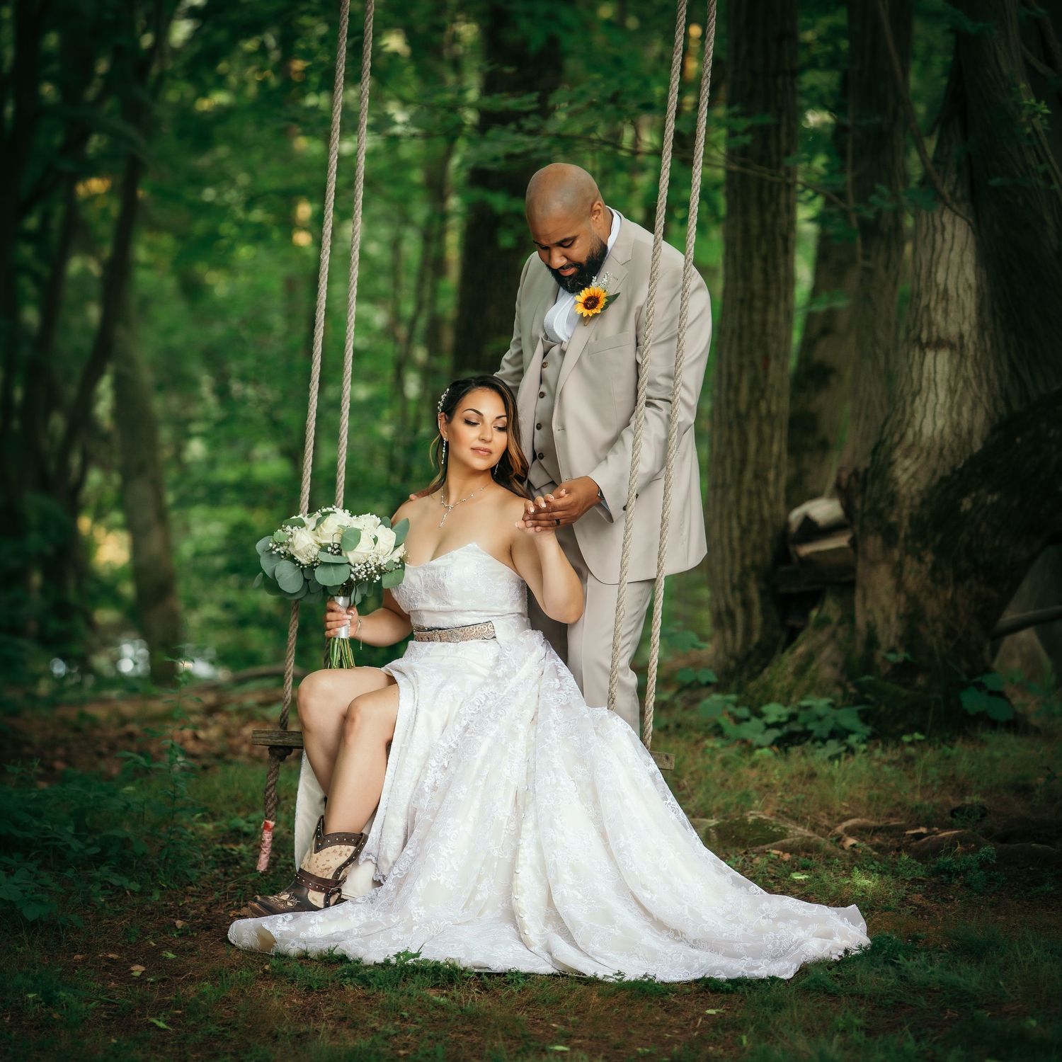 Bride in white gown sits on wooden swing as groom in tan suit stands beside her in lush green forest setting.