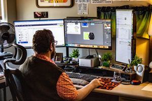 A bearded man works at a dual-monitor desktop setup, editing graphic designs in a creative home office.