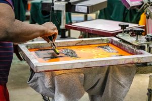 Worker applying ink through a screen printing frame onto a gray t-shirt in a professional print shop.