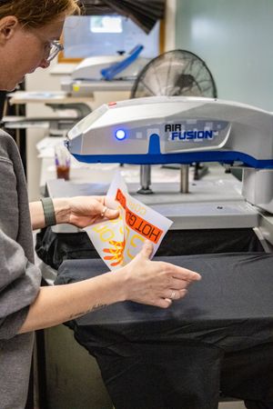 Woman peeling transfer paper from a heat press machine, applying custom graphics to dark fabric in a print shop.