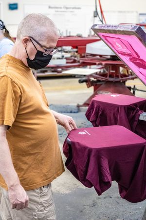 Man in black mask and orange shirt inspects maroon t-shirt on screen printing machine in workshop.