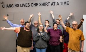 Group of people posing with arms raised under a sign reading Changing Lives One Shirt At A Time.