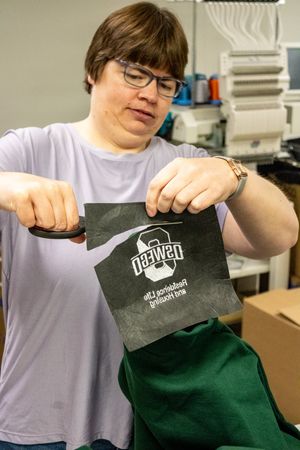 Woman holding a dark embroidered patch with a beard logo and text, working at an embroidery machine in a workshop.