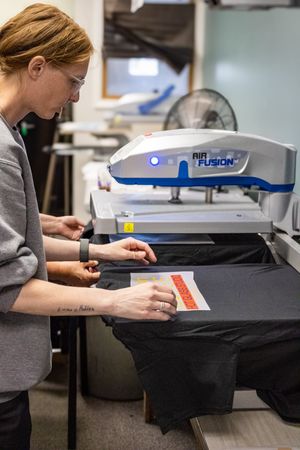 Woman using an Air Fusion heat press machine to apply a design onto a black t-shirt in a print shop.