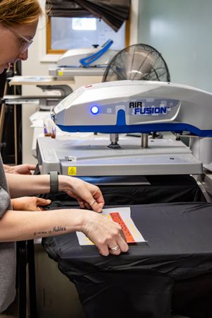 Woman using an Air Fusion heat press machine to apply a transfer onto a black garment in a print shop.