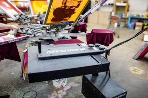 Black Flash screen printing press machine in a professional print shop with colorful screens visible in background.
