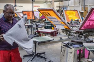 Man inspecting printed fabric in a screen printing workshop with colorful printing equipment in the background.
