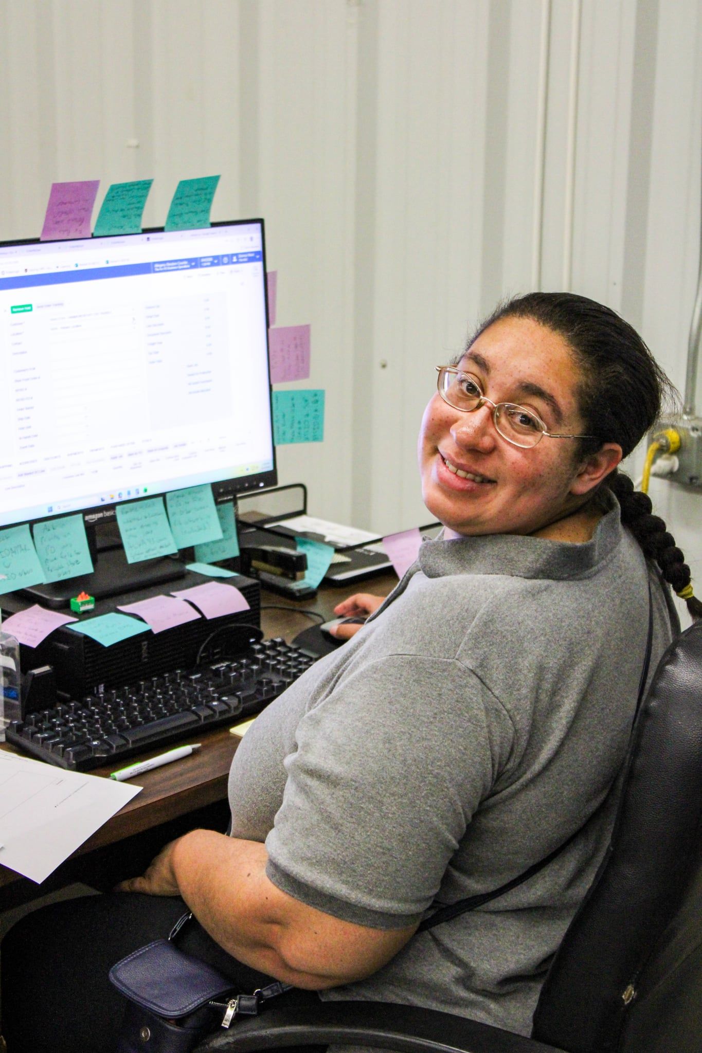 Smiling woman with glasses sitting at office desk with computer, sticky notes, and keyboard in workplace setting.