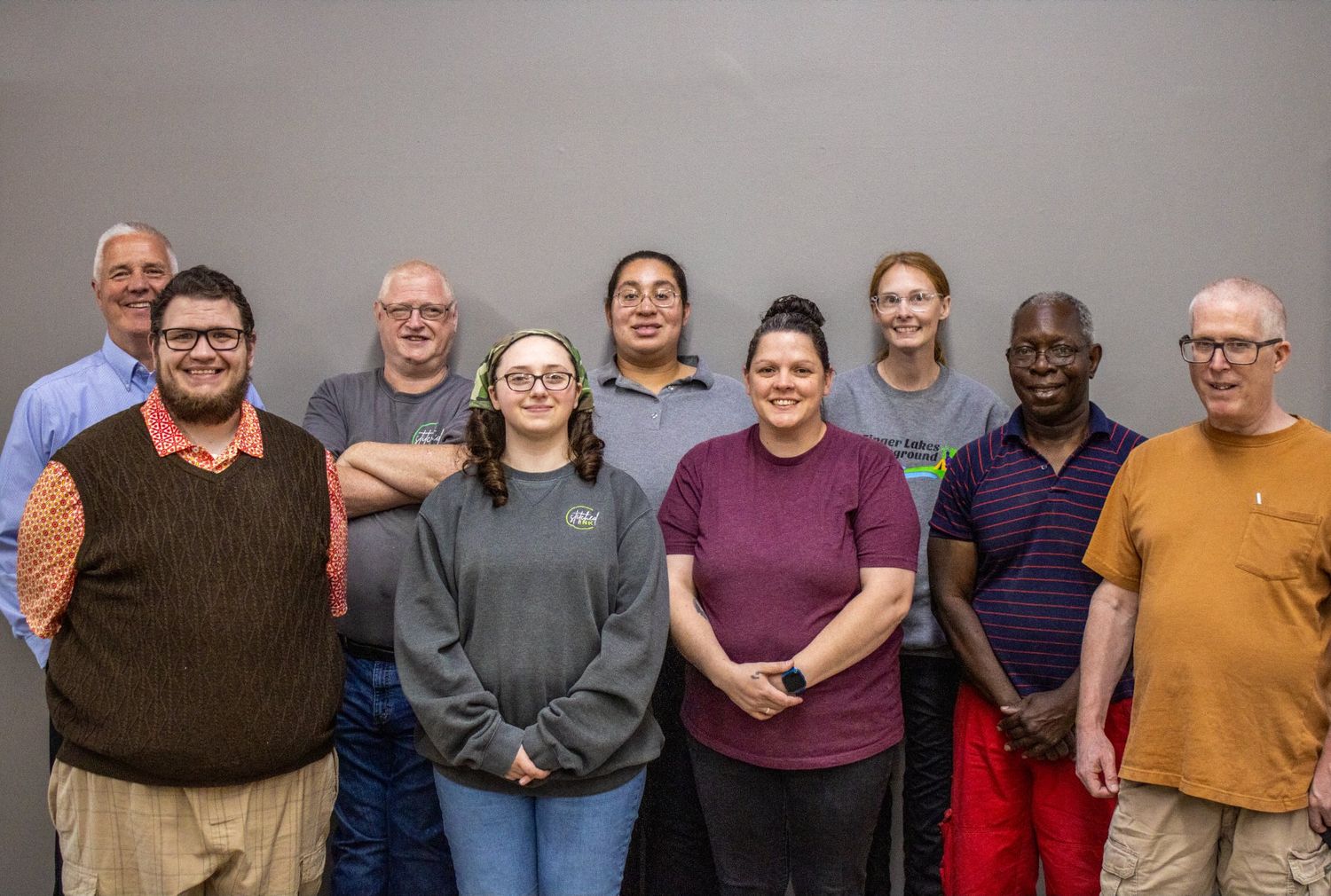 Group of nine diverse adults posing together indoors, smiling for a group photo against a gray wall.