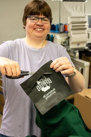 Smiling woman cutting threads on embroidered green fabric with scissors in a workshop setting.