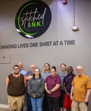 Group of employees posing in front of Stitched Ink logo wall with tagline Changing Lives One Shirt At A Time.
