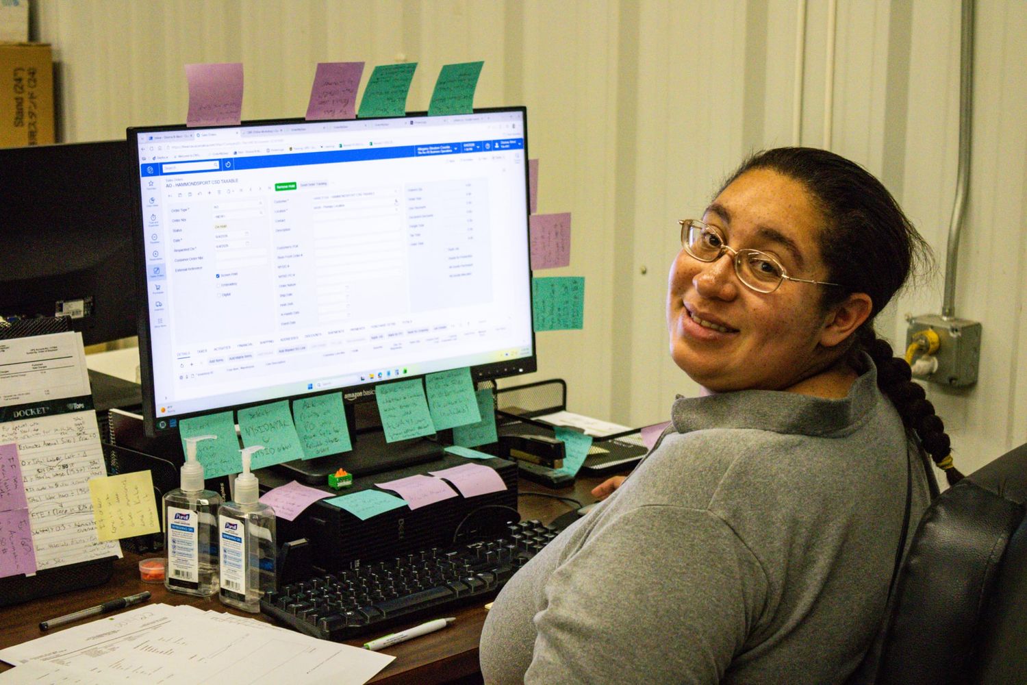 Woman smiling at desk with computer monitor, keyboard, and colorful sticky notes in an office setting.
