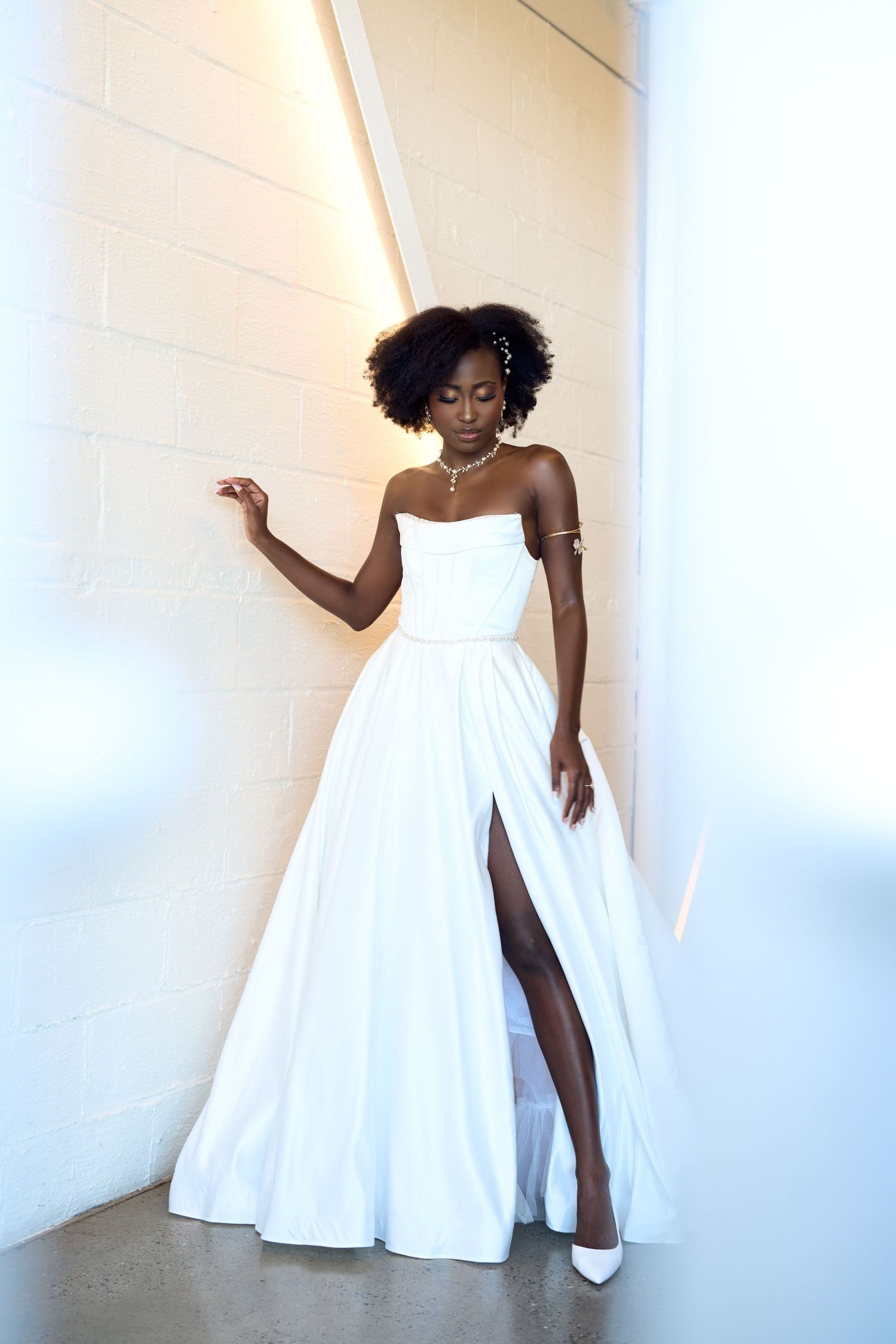 Black woman in elegant white strapless ball gown with thigh slit and heels poses gracefully against a white wall.