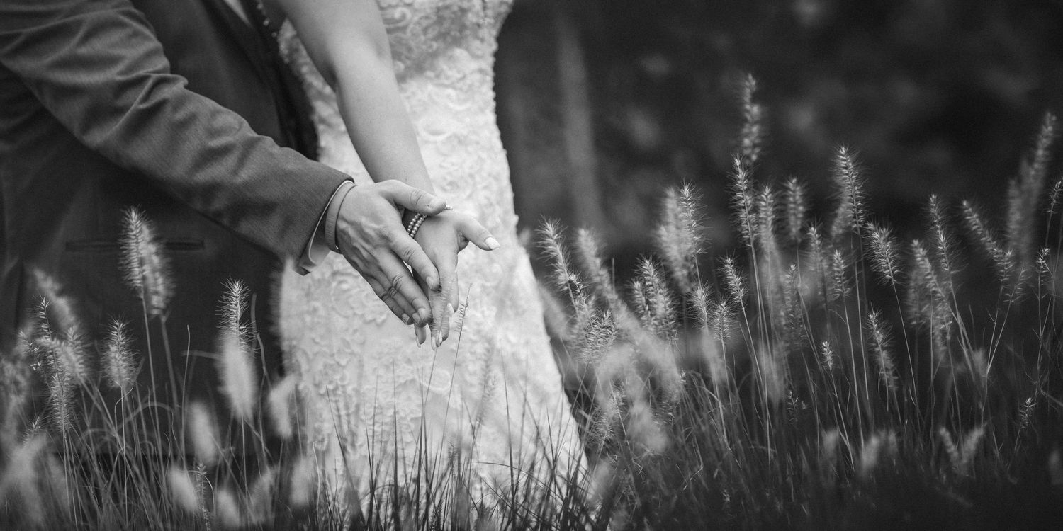 Black and white photo of a bride and groom holding hands among tall grass on their wedding day.