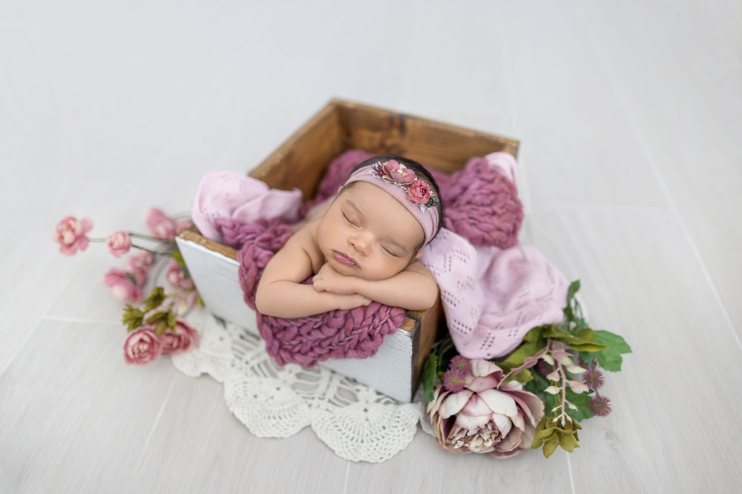 Sleeping newborn baby in wooden box with pink floral headband, surrounded by flowers and lace on white background.