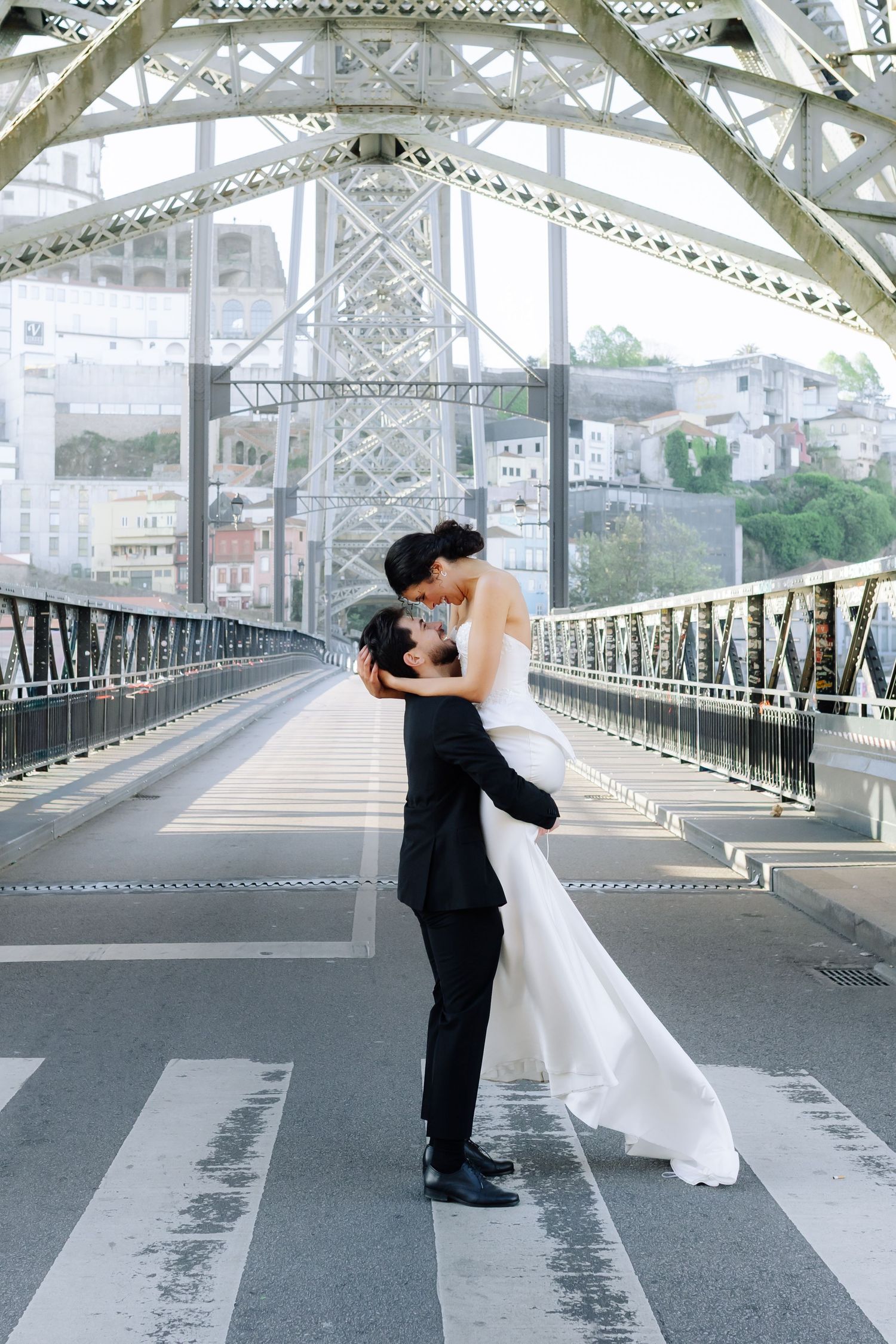 Bride and groom share a romantic kiss on a metal bridge, with the groom lifting the bride in her white wedding gown.
