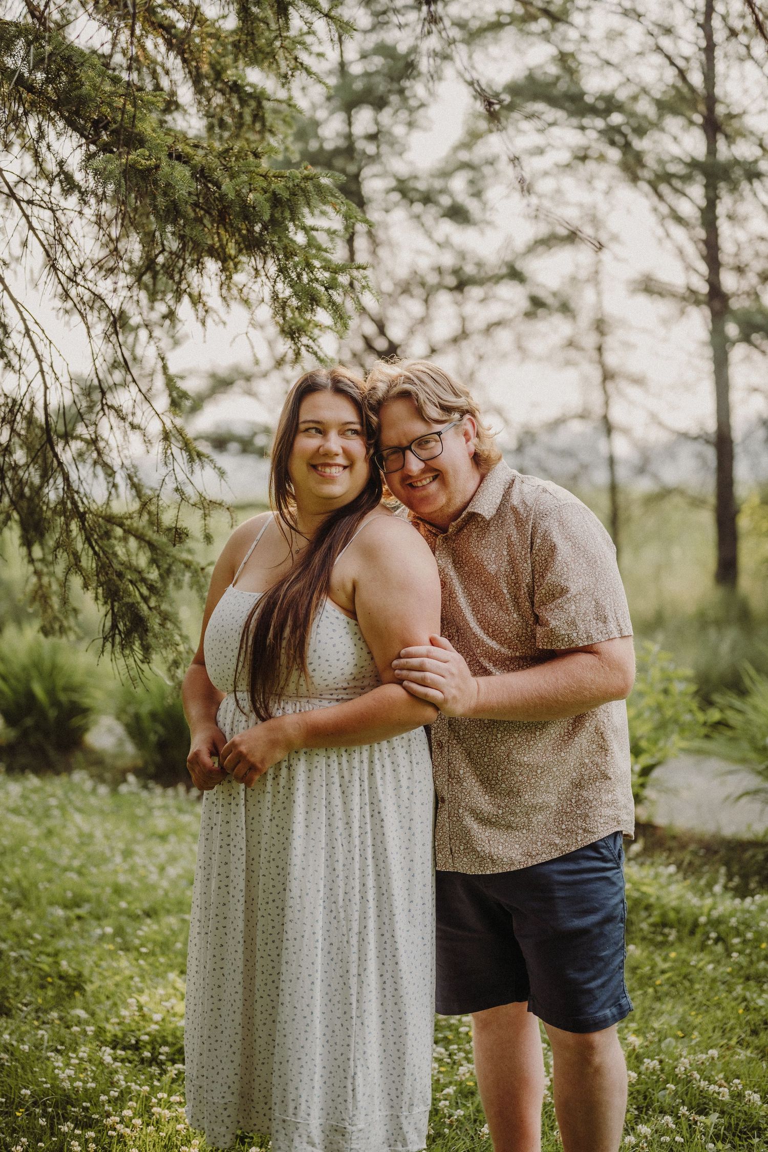 Couple sitting on birch logs in a wildflower meadow at golden hour, woman in white dress leaning against man in white shirt.