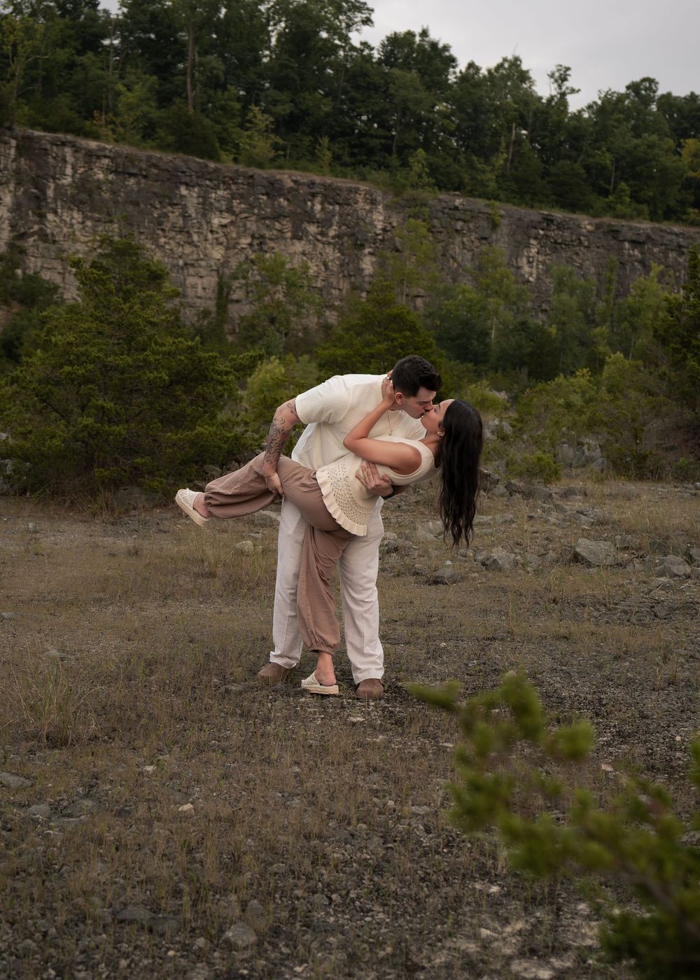 A couple shares a romantic dip kiss outdoors near a stone cliff wall surrounded by lush green trees.
