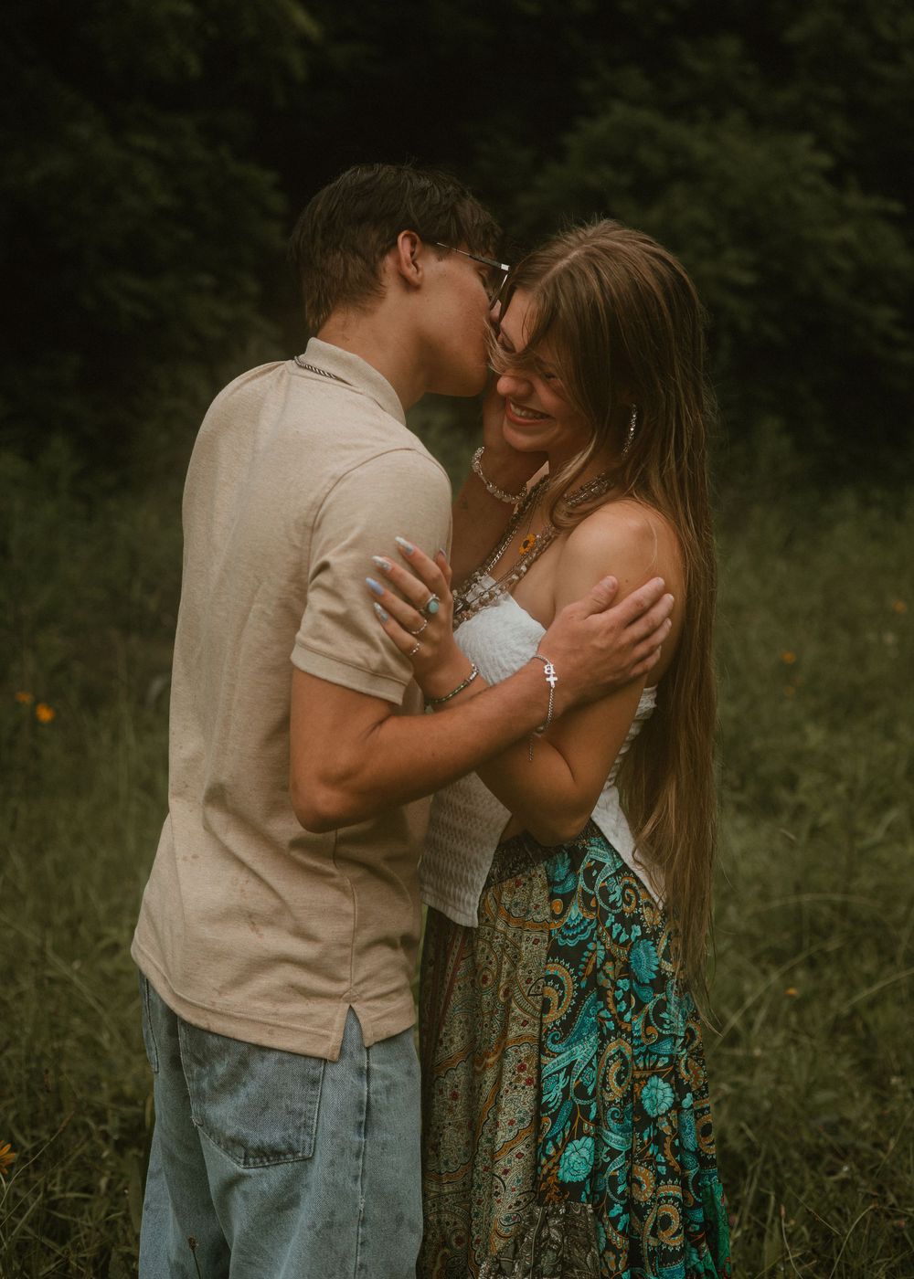 Young couple sharing a joyful kiss in a green meadow, woman in boho floral skirt laughing.