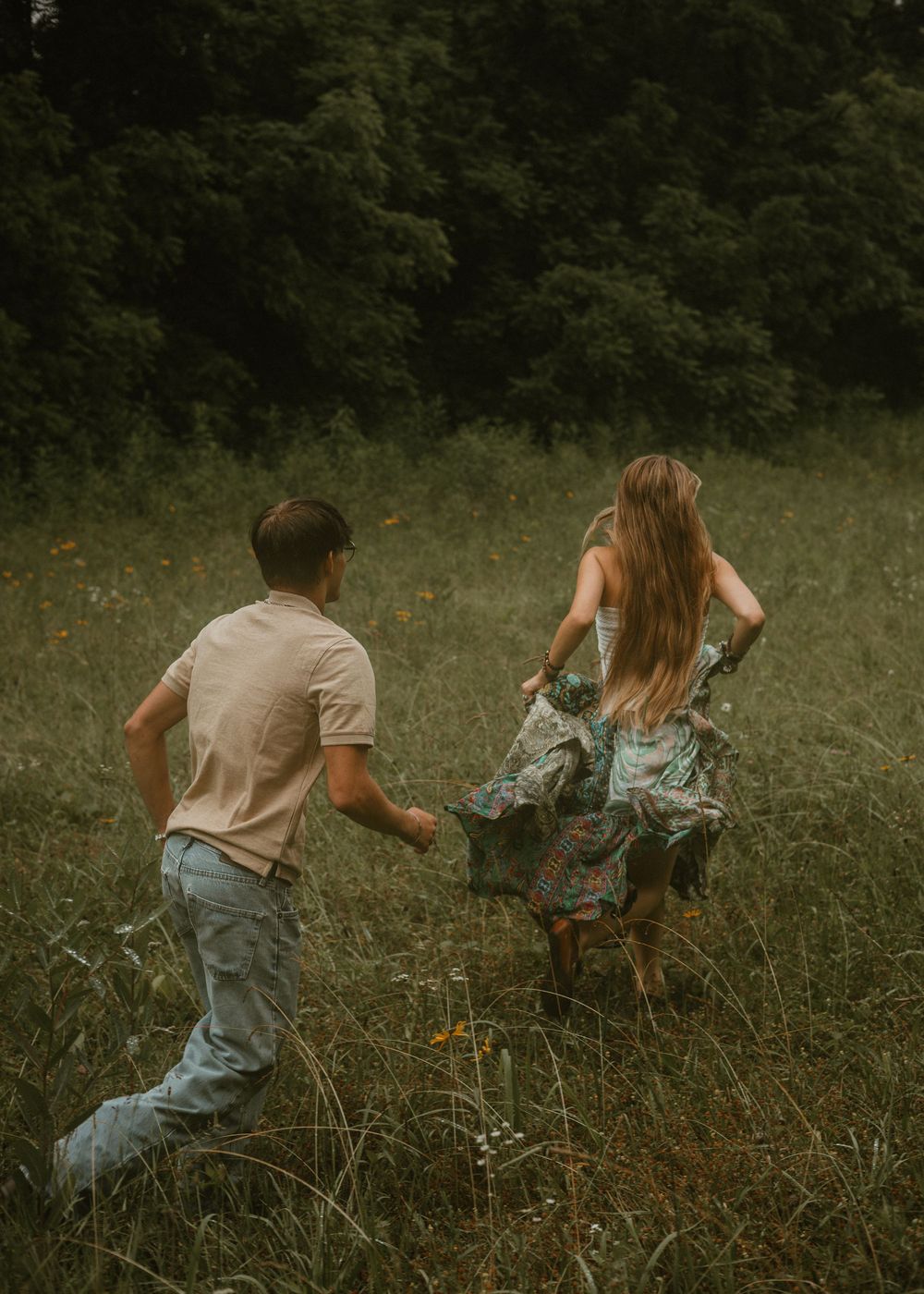 Couple running hand in hand through a wildflower meadow with lush green trees in background.