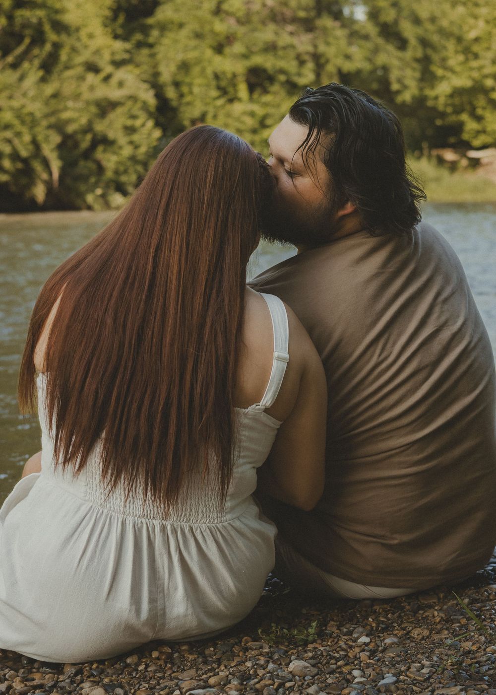 Couple sitting by a river sharing a kiss, woman with long red hair in white dress, man in brown shirt.