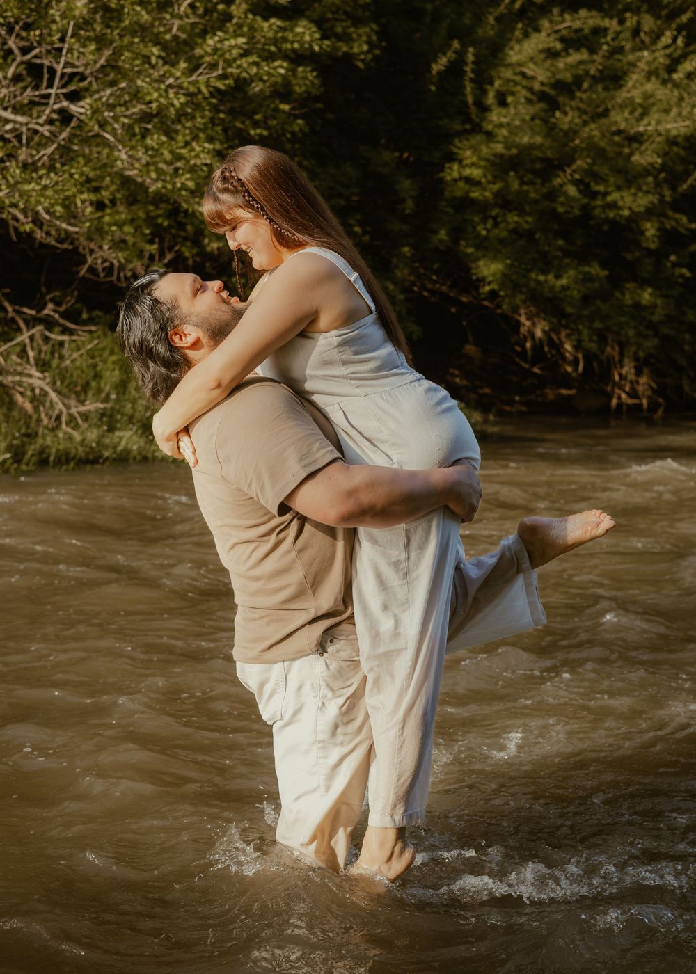 Man lifting woman in his arms while standing in a sunlit river, both laughing joyfully.