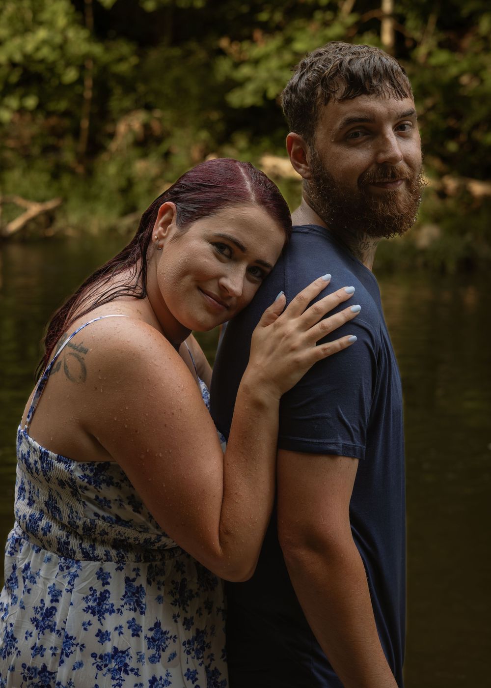 Smiling woman with purple hair leans on bearded mans shoulder near a serene woodland creek.