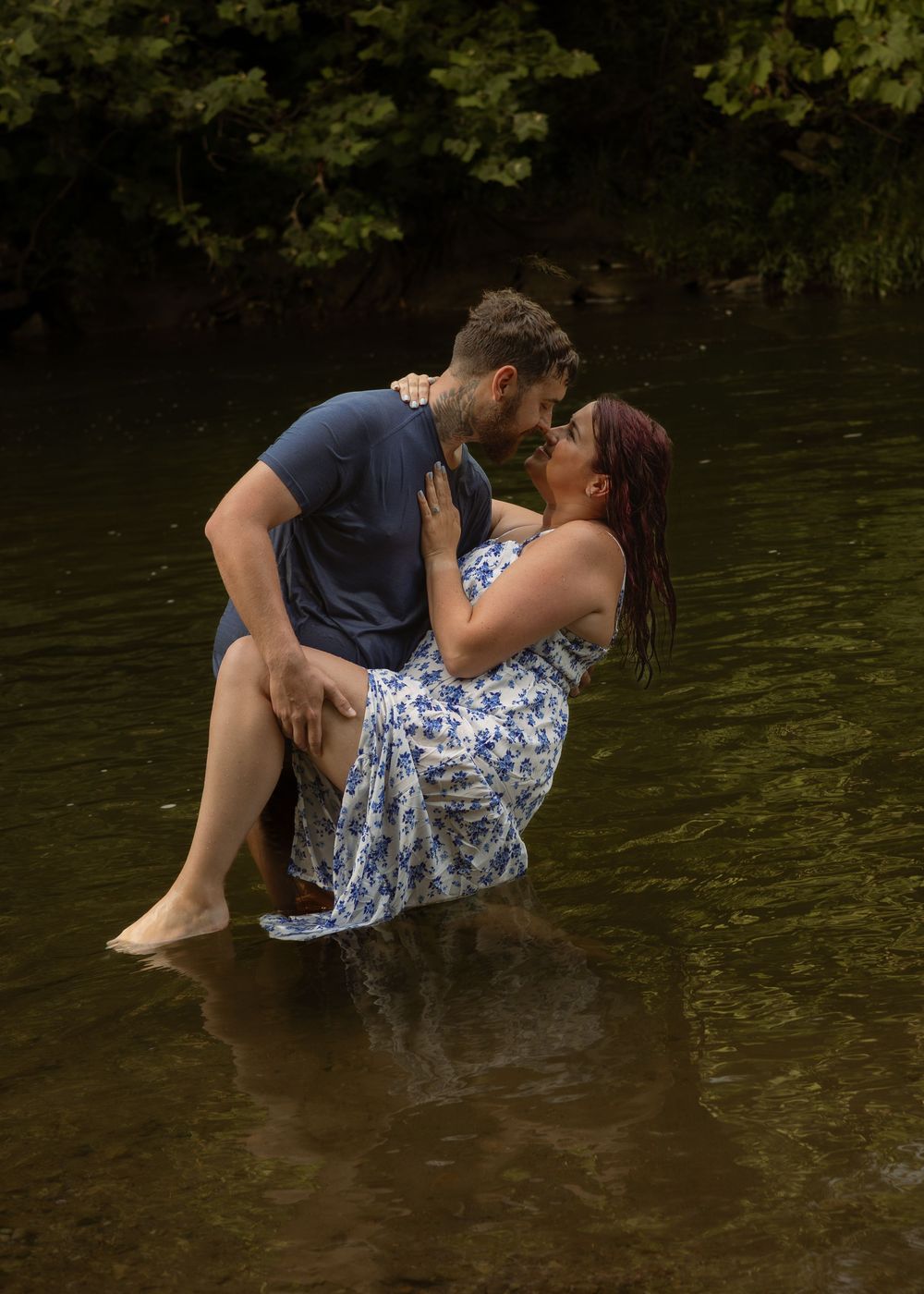 Man holding woman in his arms while standing in a shallow creek surrounded by lush greenery.